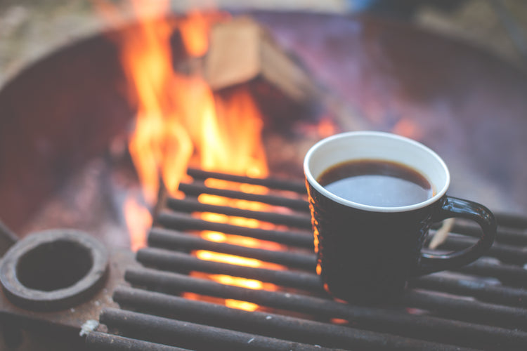 A campfire with with a cooking grate and a steaming hot cup of coffee on it.